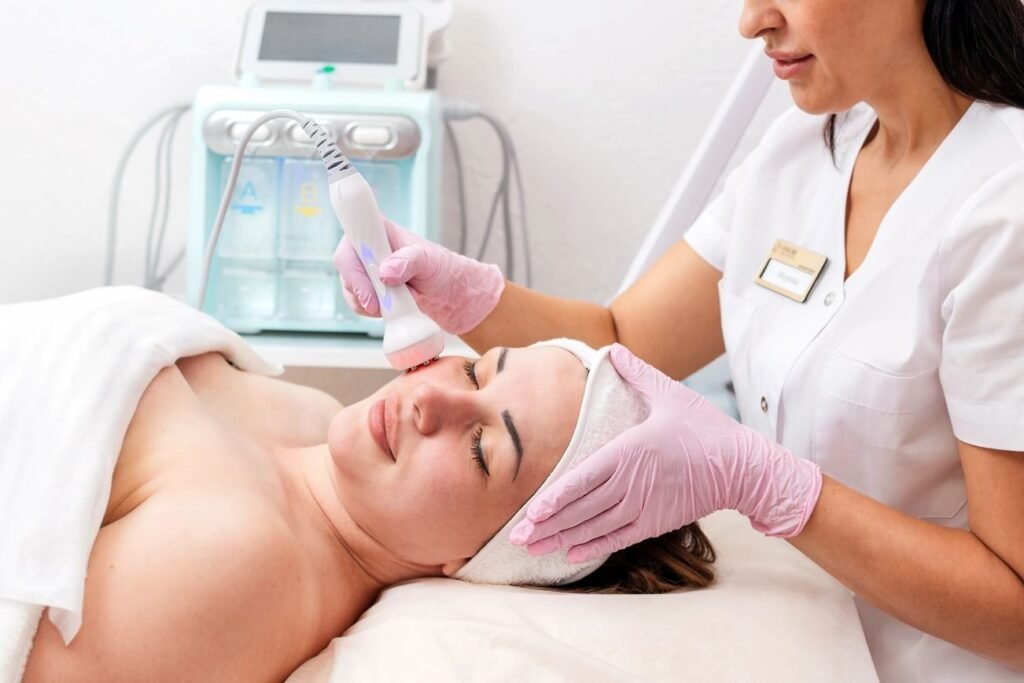 Aesthetician performing Fractional Radio Frequency (RF) treatment on a relaxed woman in a spa setting. The patient is lying on a treatment bed with a towel wrapped around her head.