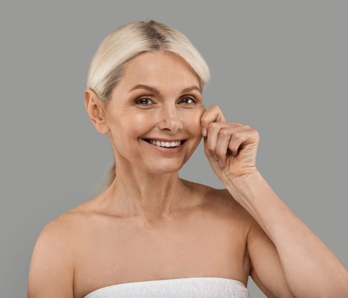 Pico Laser Treatment | Laser Face Rejuvenation 8 Smiling mature woman with firm, youthful skin pinching her cheek to showcase skin elasticity, representing the benefits of Sublime Skin Firming treatment.